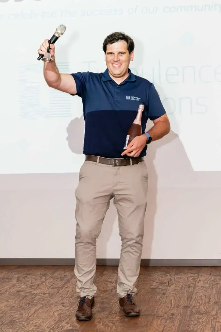 Mann in Poloshirt hält Mikrofon und Champagnerflasche bei Firmevent Wien auf der Bühne