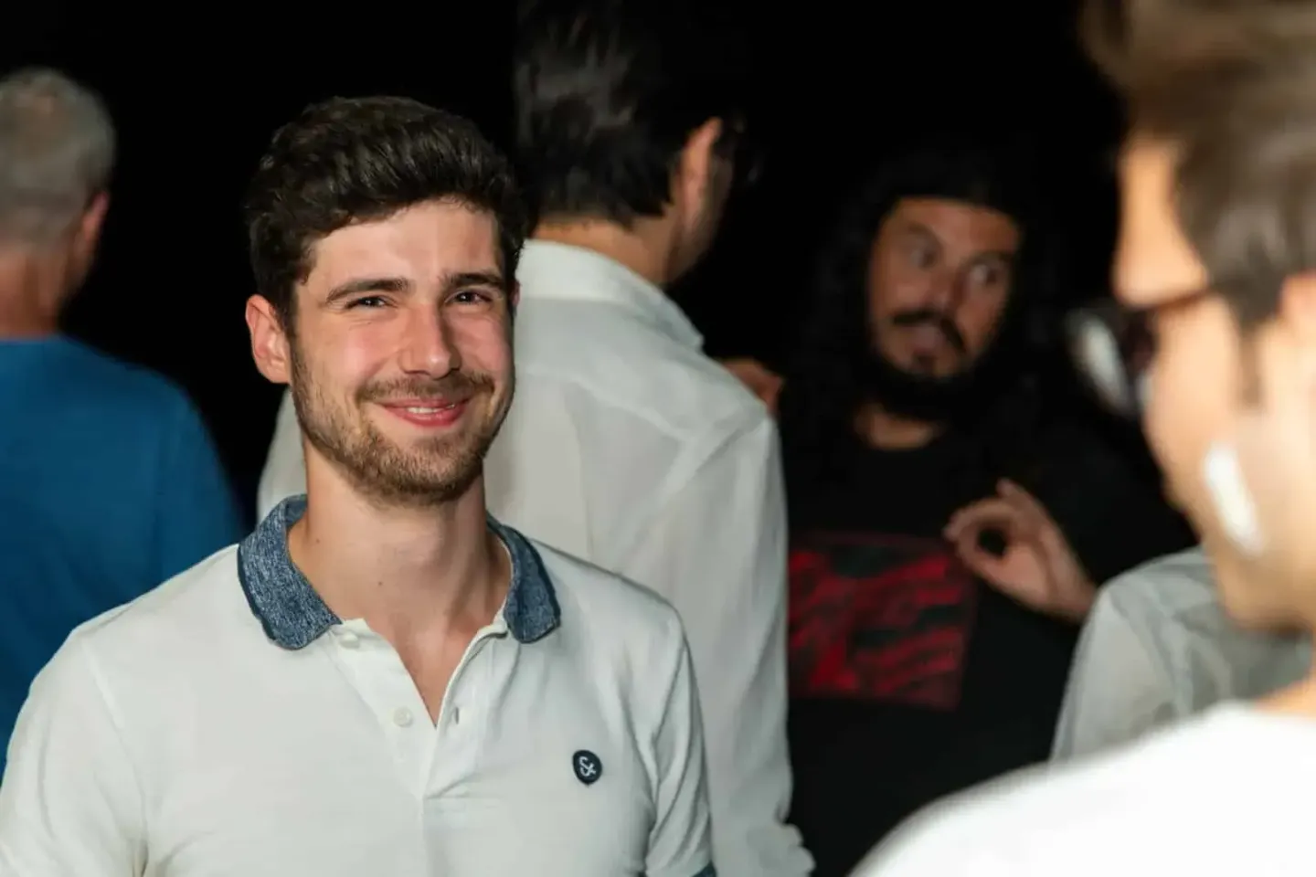 Lächelnder Mann in weißem Poloshirt mit Denim-Kragen bei einem Firmenevent in Wien, umgeben von Gästen