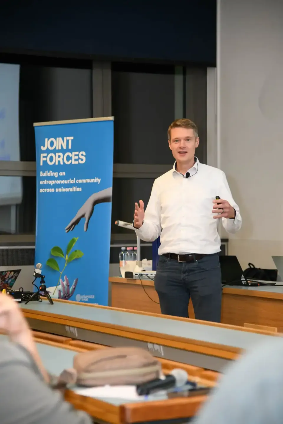 Junger Mann präsentiert beim Joint Forces Event an der TU Wien vor blauem Banner mit Publikum im Hörsaal