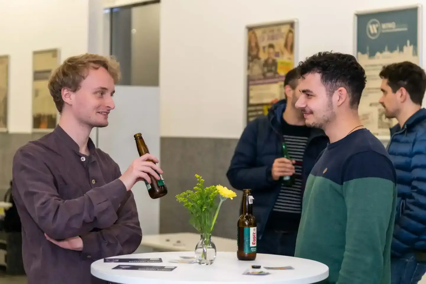 Junge Männer im Gespräch bei einem Firmenevent in Wien, einer hält eine Bierflasche, gelbe Blumen auf dem Tisch