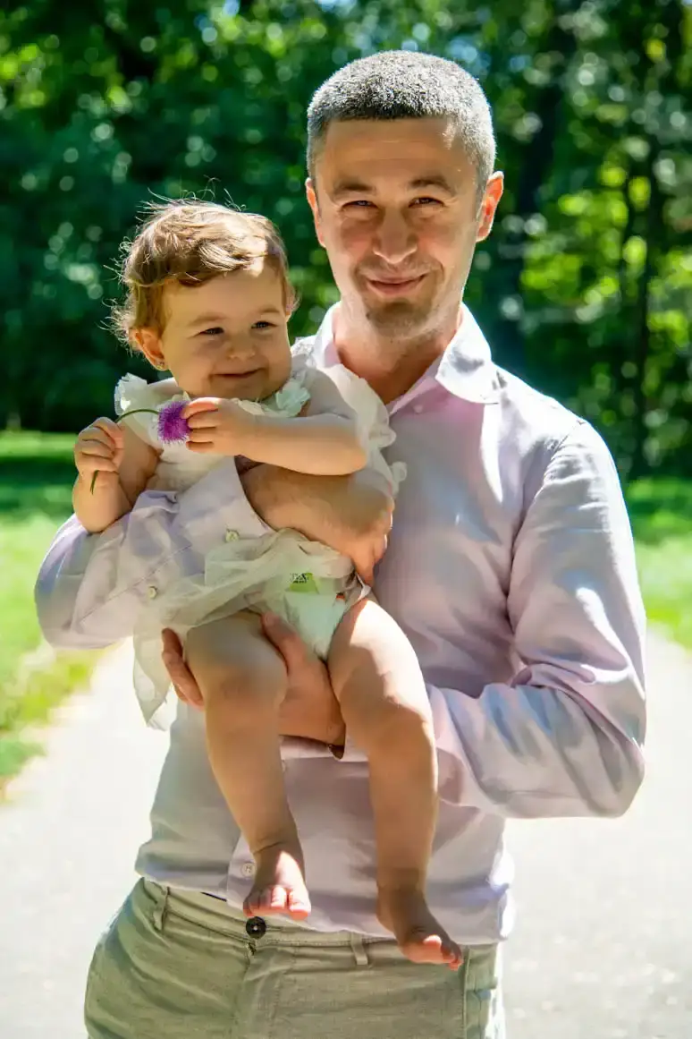 Vater hält lächelndes Kleinkind mit Blume im Arm, Familienfotografie Wien, sonniger Garten im Hintergrund