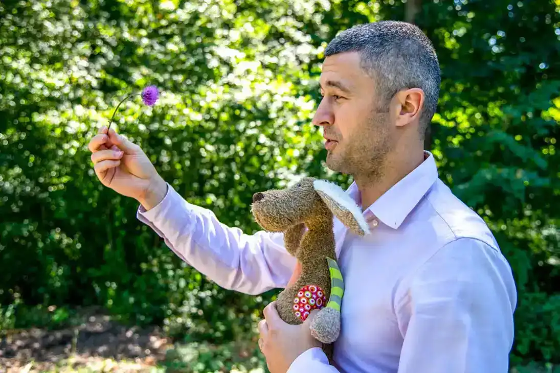 Mann mit Teddy in der Hand hält eine lila Blume, Familienfotos Wien mit grünem Gartenhintergrund