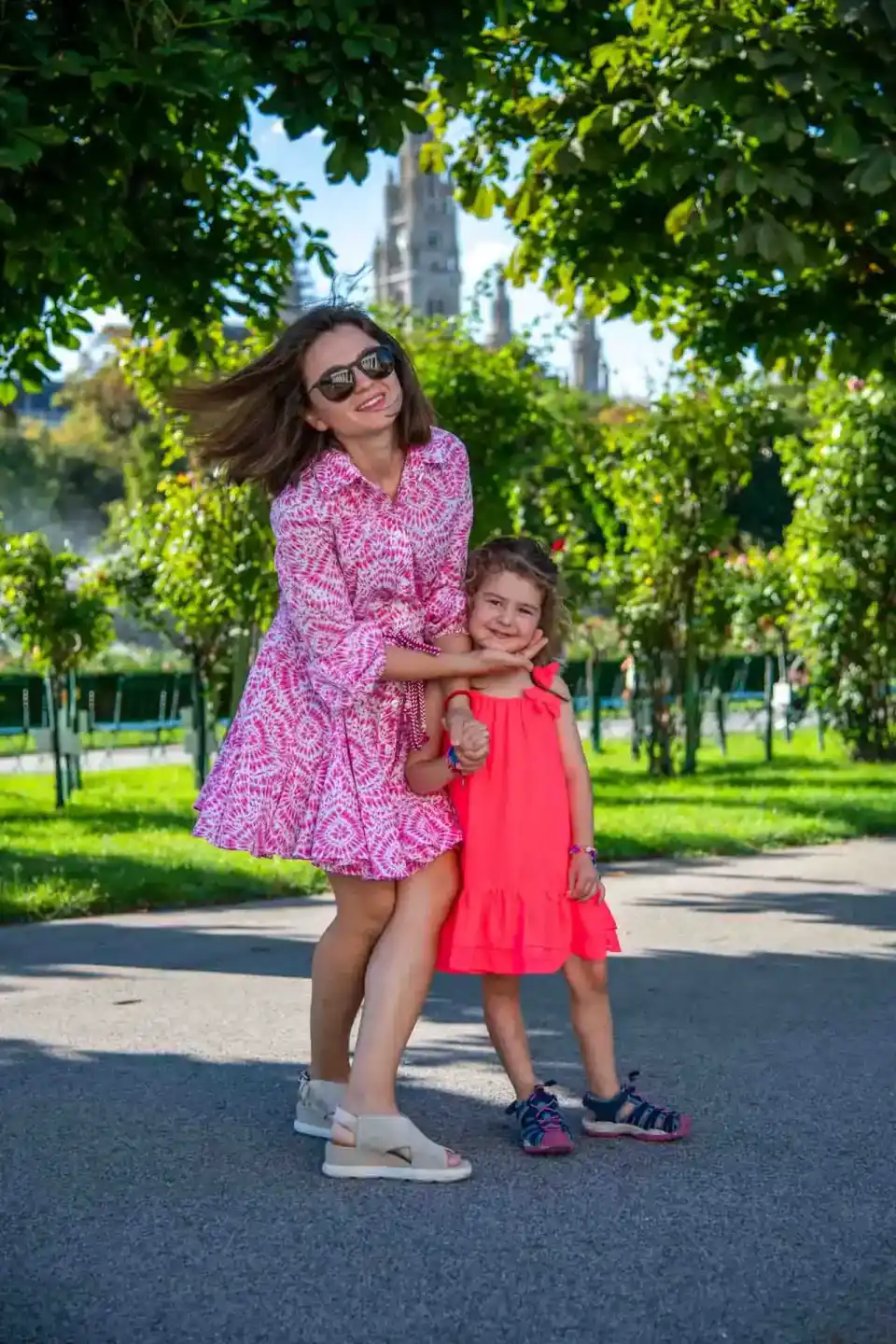 Mutter und Tochter in farbenfrohen Kleidern posieren lächelnd in einer grünen Parkanlage mit Kirche im Hintergrund - Familien