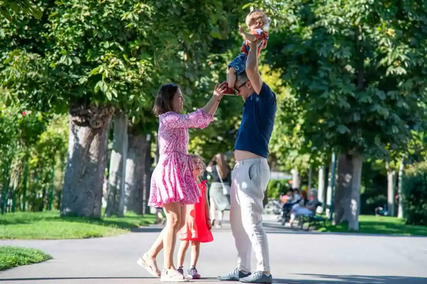Vater wirft lachendes Kleinkind in die Luft während Mutter zuschaut in einem Park – Familienfotografie Wien