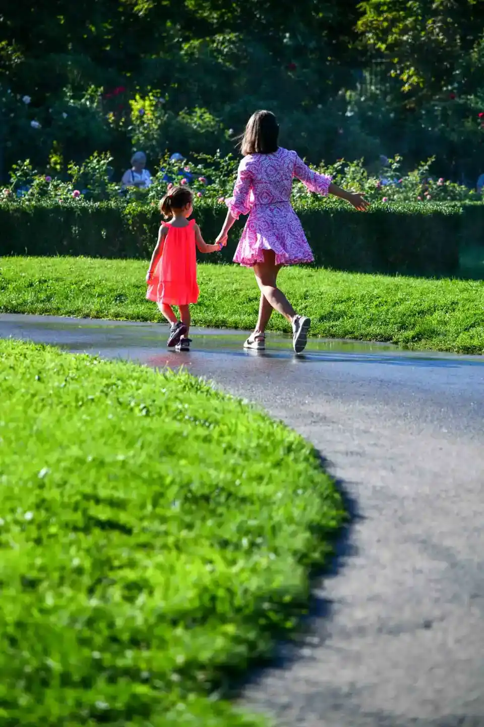 Mutter und Tochter laufen Hand in Hand auf einer nassen Gartenpathode, umgeben von grünen Hecken und bunten Blumen – professi
