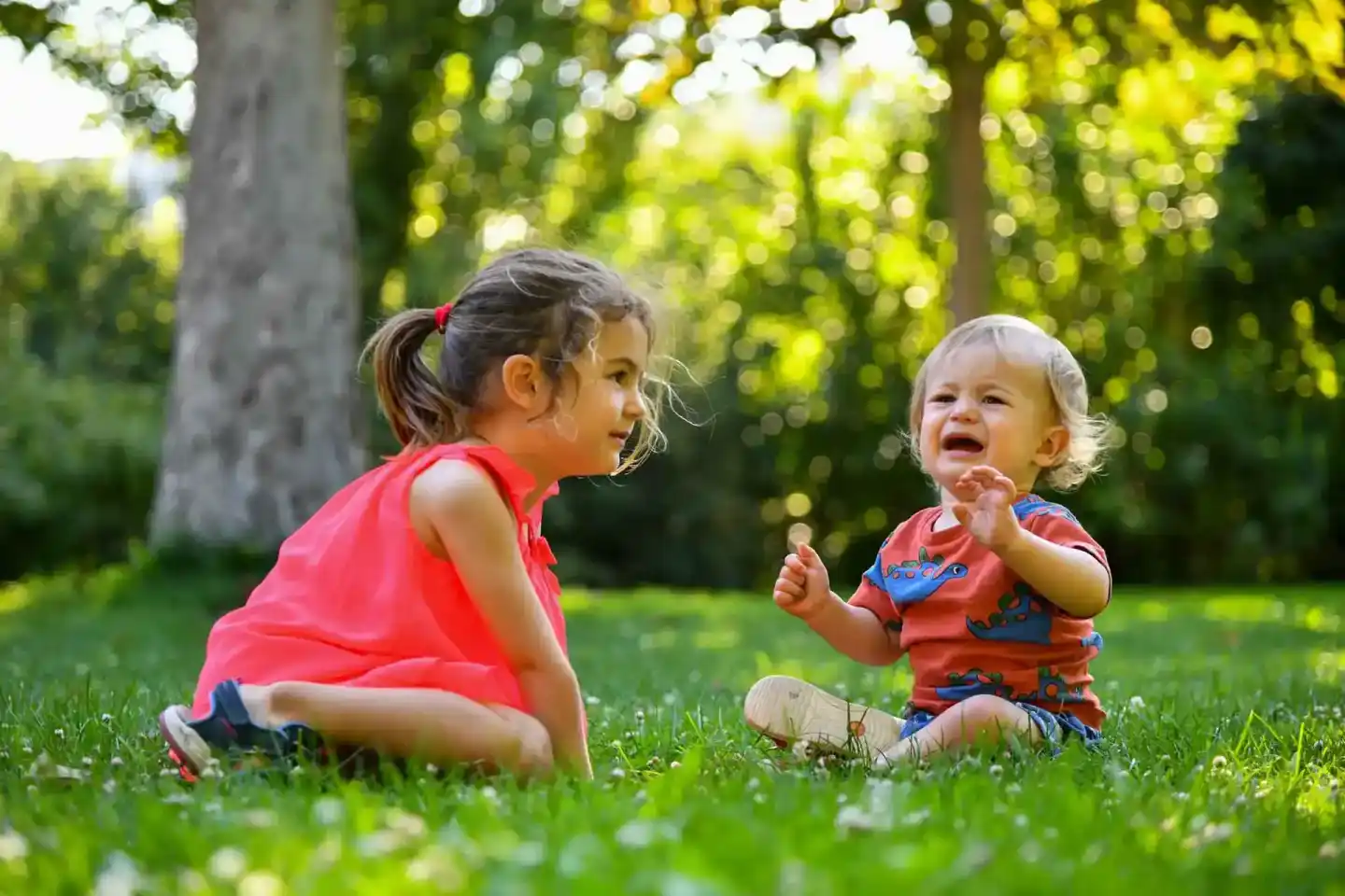 Zwei kleine Kinder sitzen lachend auf einer grünen Wiese unter Bäumen – professionelle Familienfotografie Wien