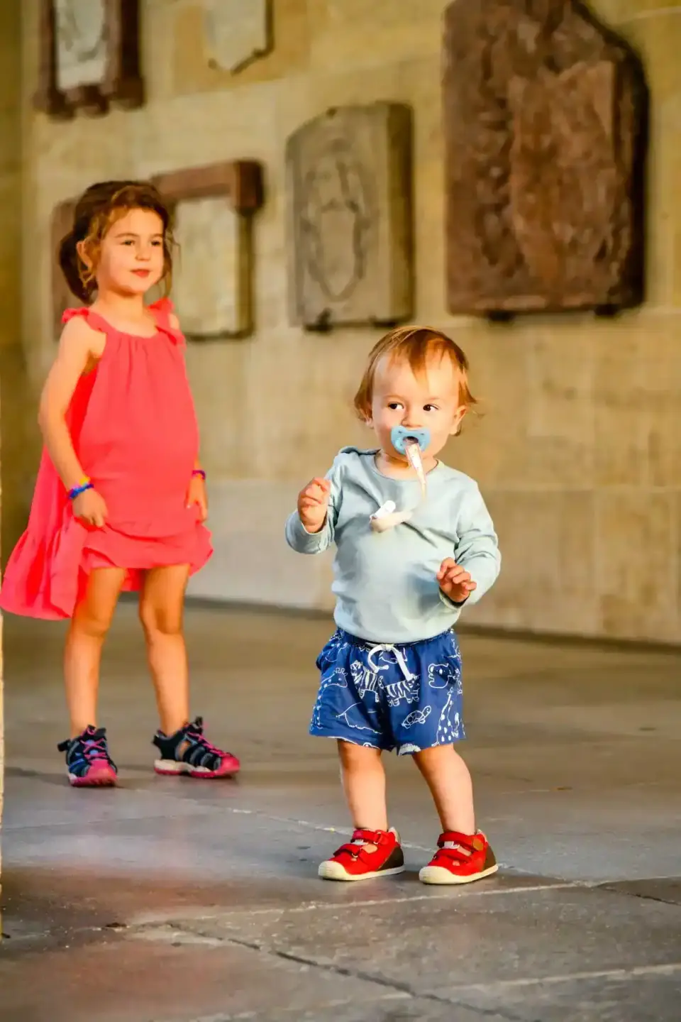 Zwei kleine Kinder spielen in einem historischen Innenhof: Ein Mädchen in rosa Kleid und ein Junge in blauem Outfit mit Lolly