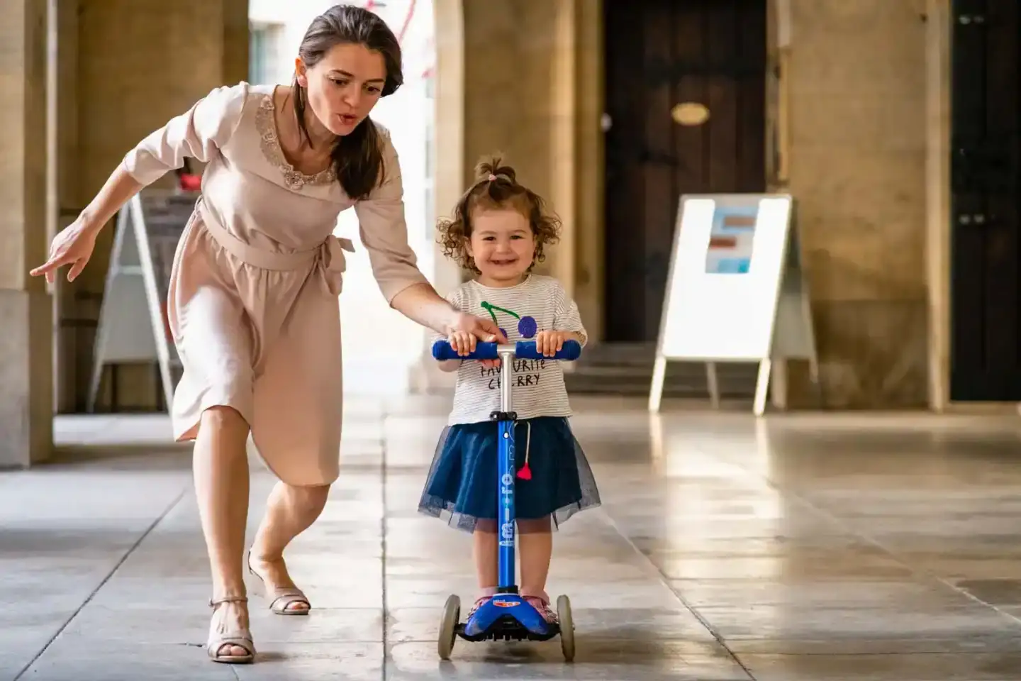 Mutter läuft mit ihrer Tochter auf einem Roller durch einen hellen Flur - Familienfotografie Wien