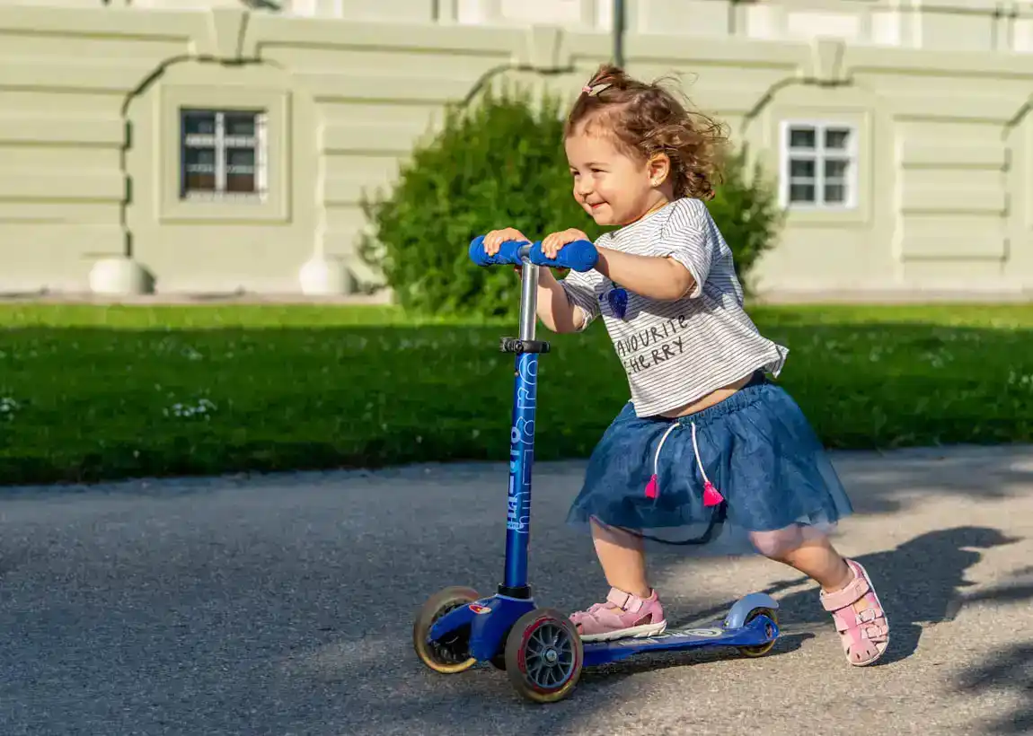 Kleines Mädchen mit lockigem Haar fährt lächelnd Roller vor einem hellen Haus – Familienfotografie Wien