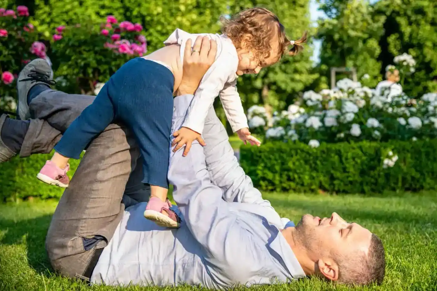 Vater spielt mit kleinem Kind im sonnigen Garten, Familienfotografie Wien mit blühenden Blumen im Hintergrund