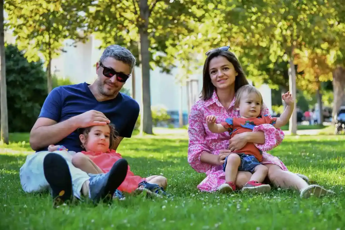 Familie mit zwei Kleinkindern sitzt auf einer grünen Wiese unter Bäumen. Professionelle Familienfotografie Wien.
