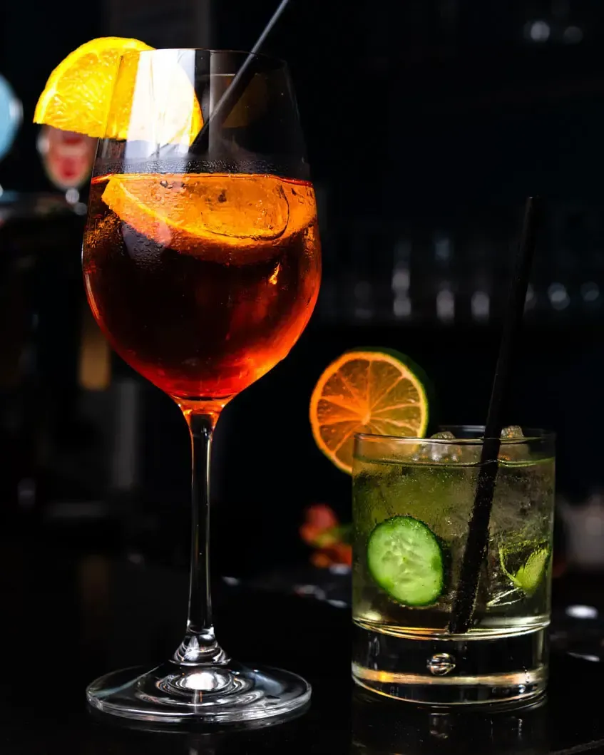 Zwei elegante Cocktails auf schwarzem Hintergrund: ein rotes Aperitif-Getränk mit Orangenscheibe und ein grüner Mojito mit Gu