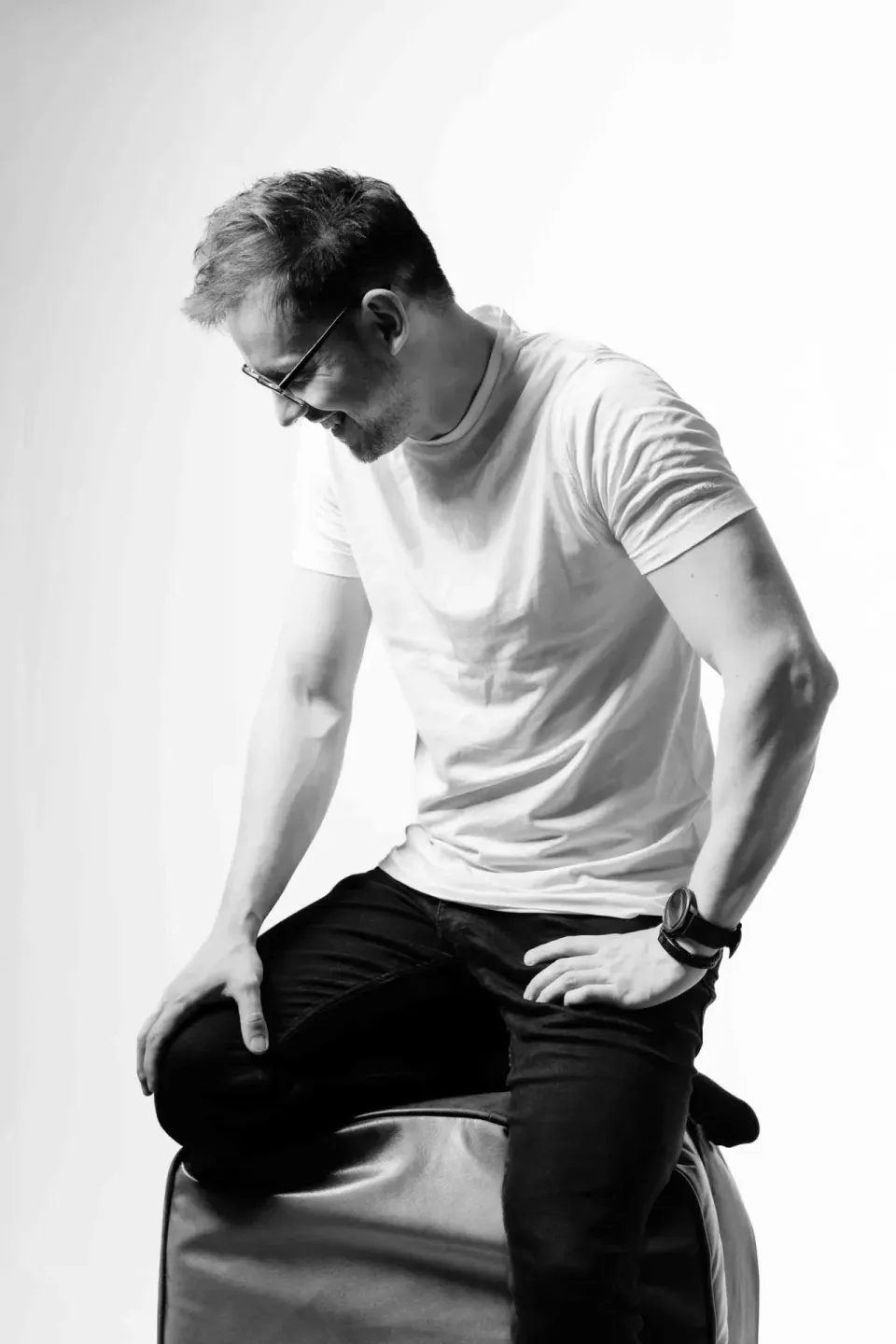 Mann mit Brille in weißem T-Shirt und dunkler Jeans sitzt auf einem Hocker, blickt nach unten - professionelles Portrait Wien