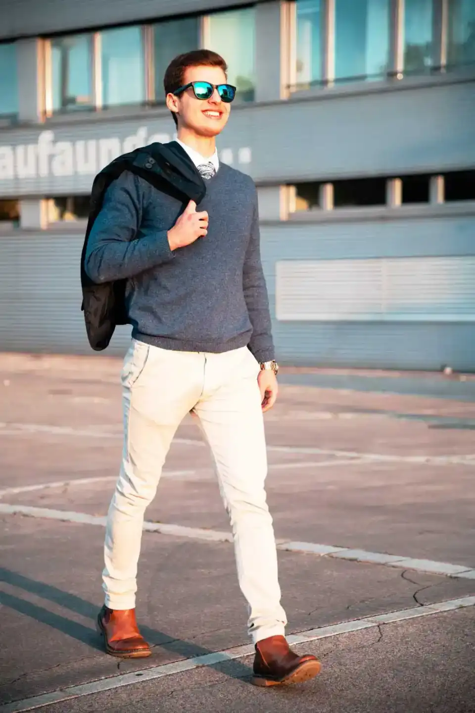 Junger Mann in grauem Pullover und weißer Hose mit Sonnenbrille vor modernem Gebäude - Portrait Wien