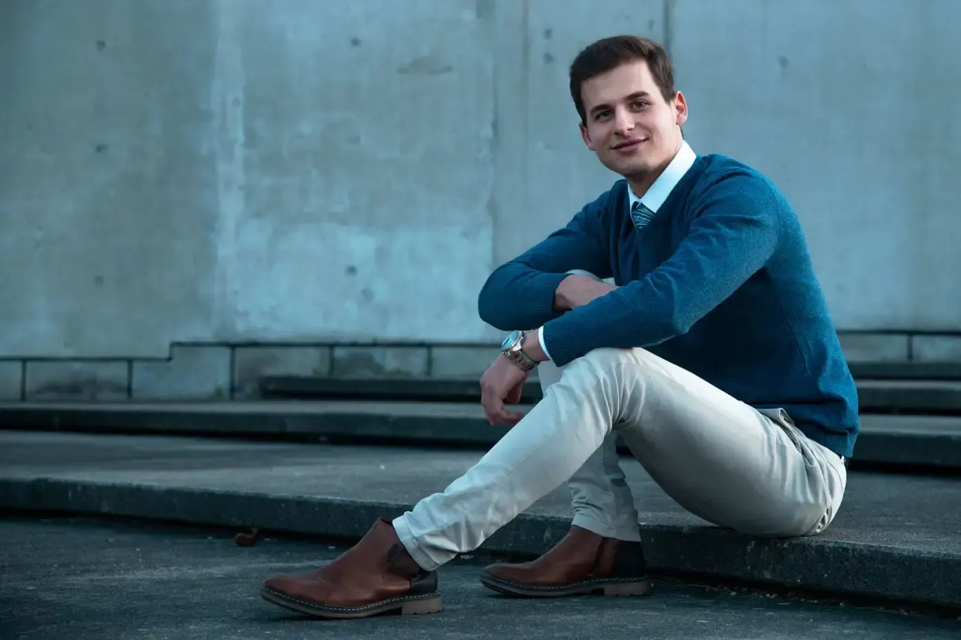 Junger Mann in blauem Pullover und weißer Hose sitzt vor grauer Betonwand - professionelles Portrait Wien
