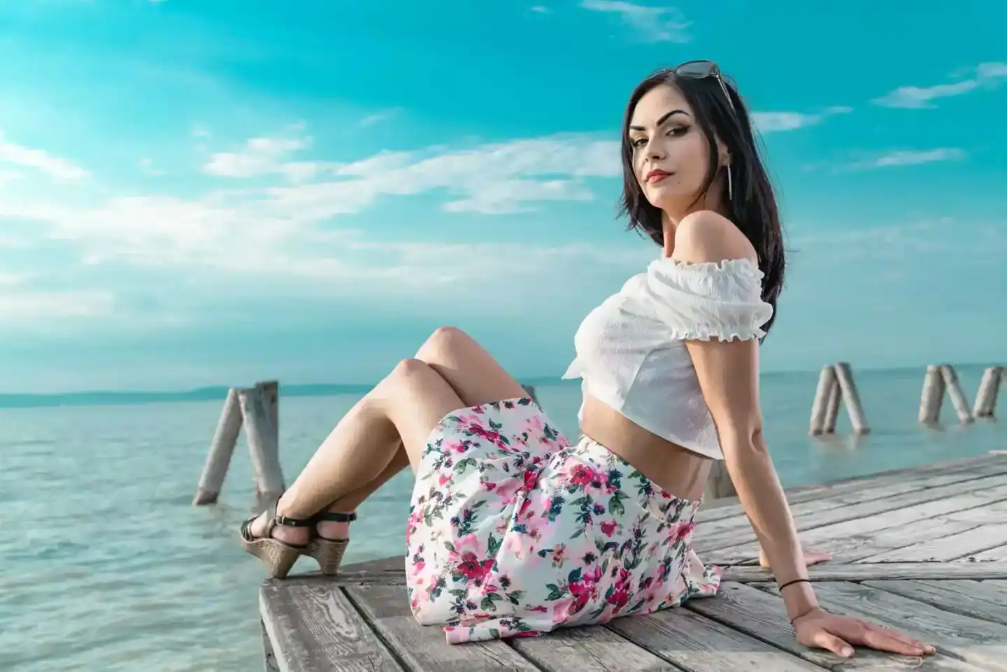 Junge Frau in weißem Crop Top und floralem Rock sitzt auf einem Holzsteg am türkisblauen Meer – professionelles Portrait Foto