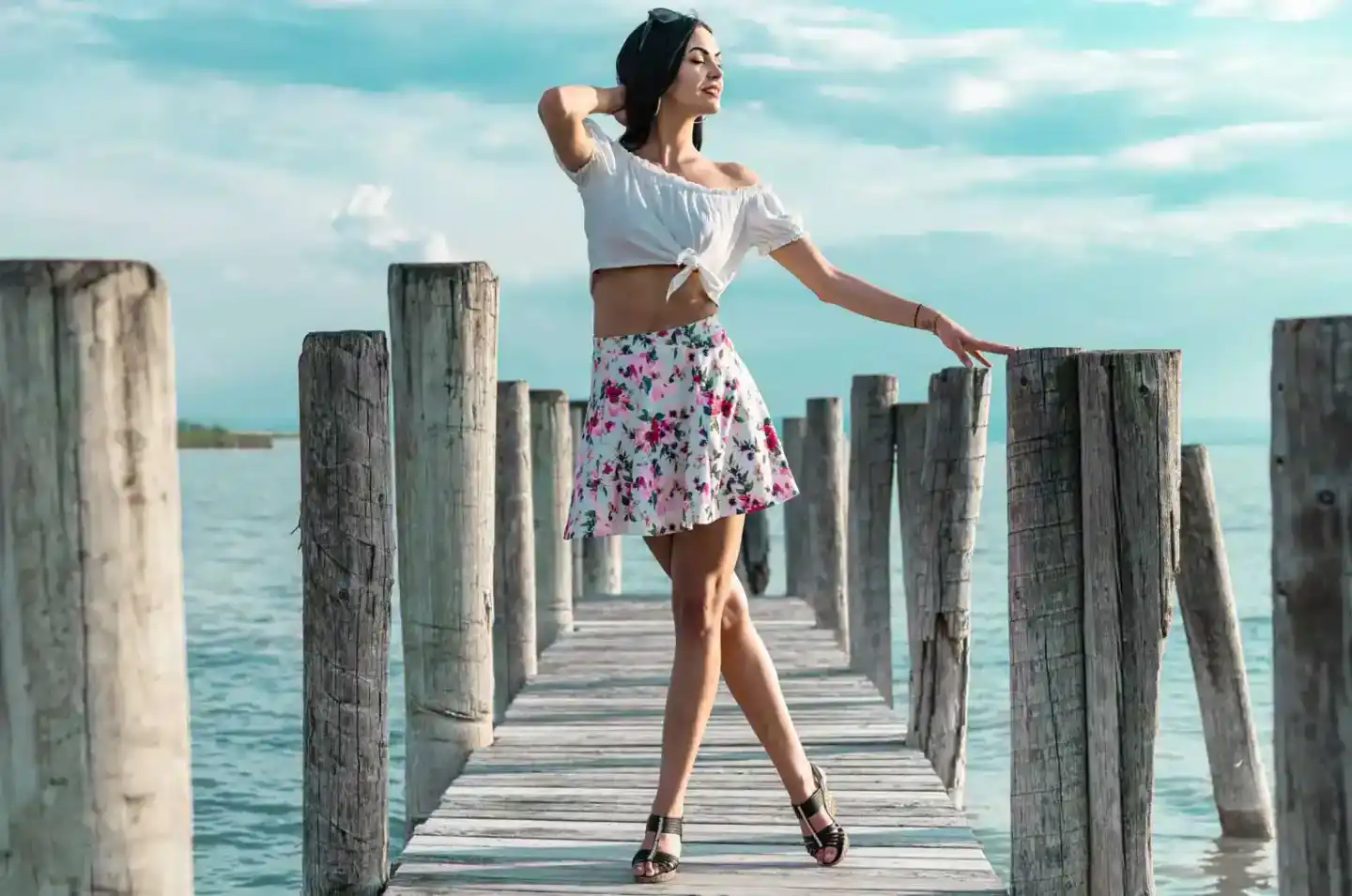 Elegante Frau in weißem Crop Top und Blumenrock posiert auf einem Holzsteg am türkisblauen Meer für professionelle Portraitfo