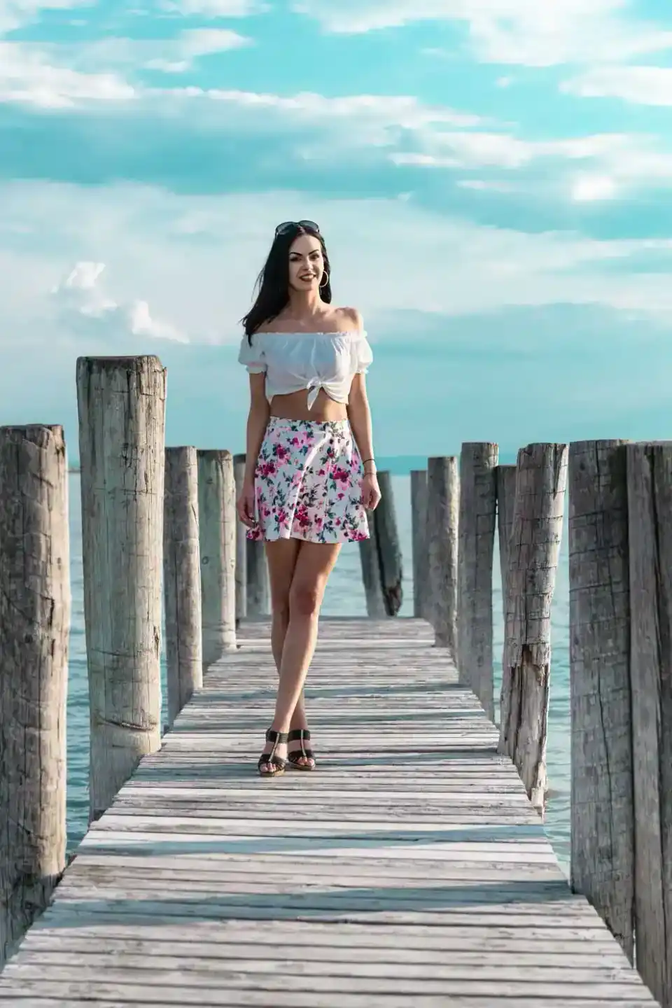 Junge Frau in weißem Crop Top und Blumenmuster-Rock auf einem Holzsteg am Meer posiert für Portrait Wien