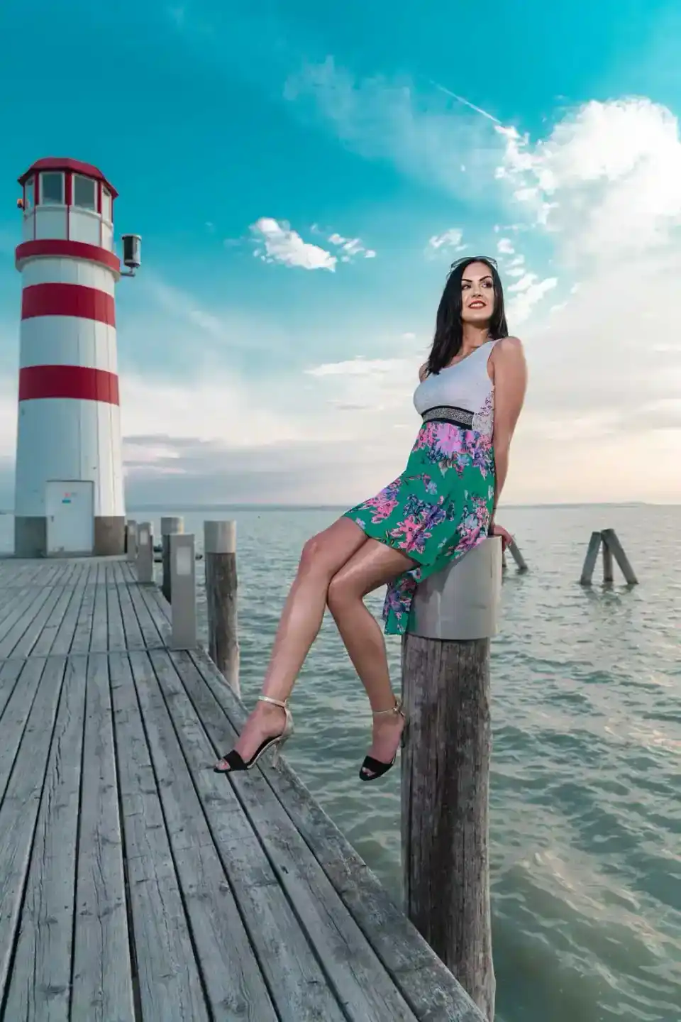 Frau in farbenfrohem Kleid sitzt auf Holzpfosten eines Stegs vor rot-weiß gestreiftem Leuchtturm am Wasser – professionelles 