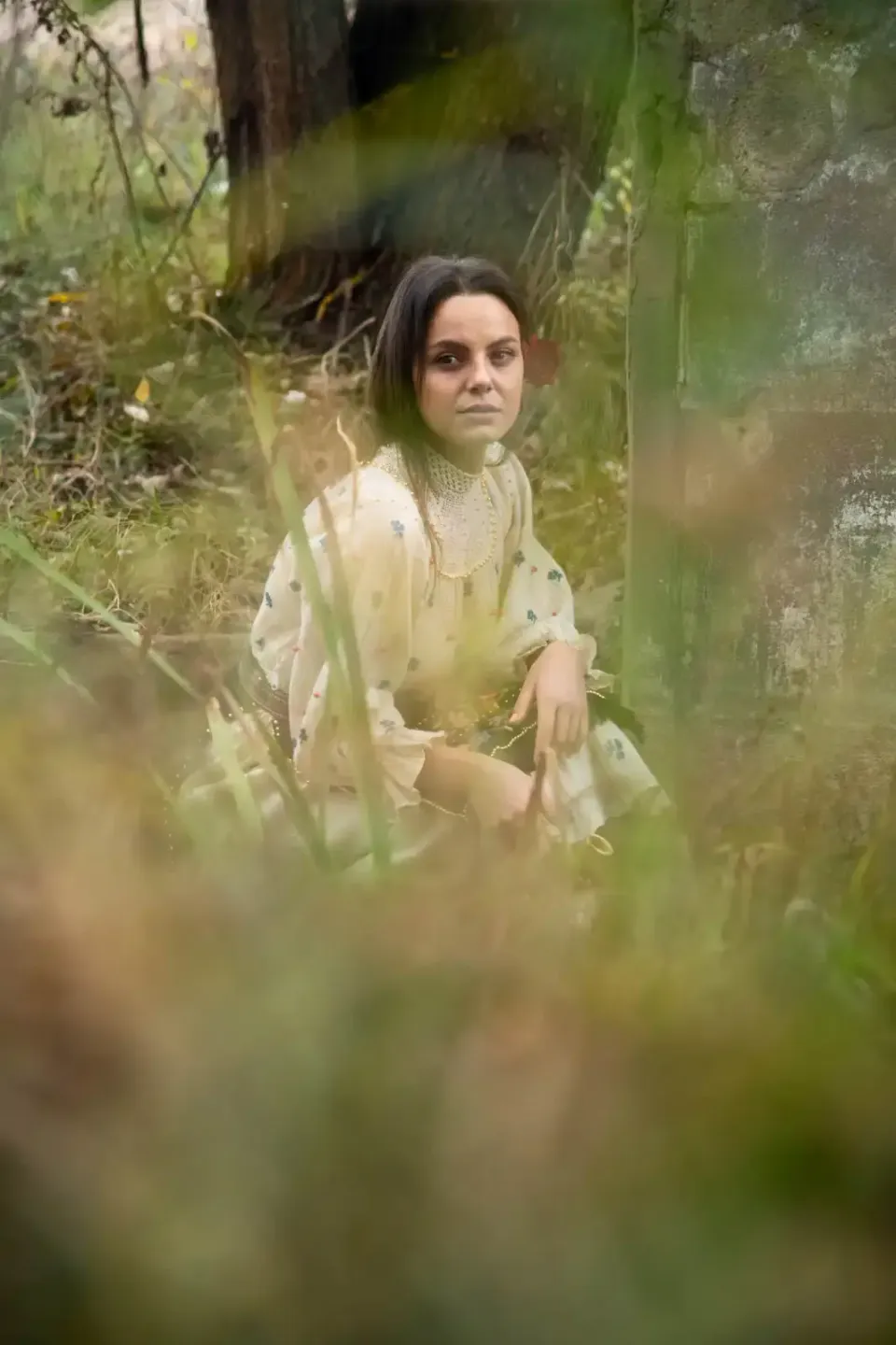 Junge Frau in cremefarbenem Leinenhemd sitzt in verwildertem Garten zwischen Pflanzen, Portrait Wien