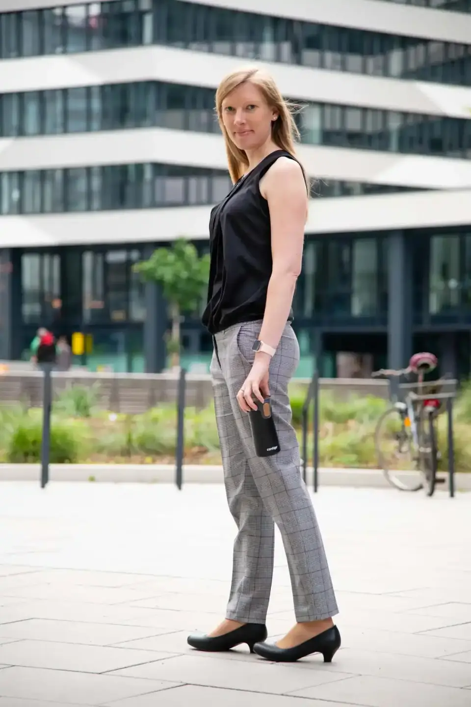 Junge Frau in schwarzem Oberteil und graukarierter Hose steht selbstbewusst vor modernem Gebäude – professionelles Portrait W