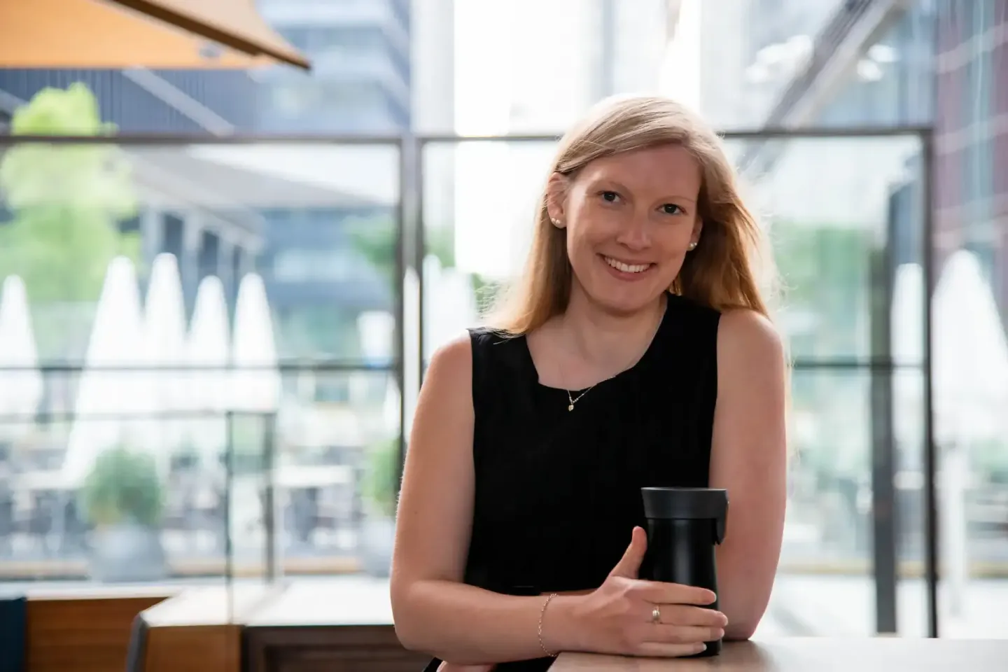Portrait einer lächelnden Frau mit rötlichblondem Haar in schwarzem Oberteil vor modernem Glasgebäude mit Terrasse - Portrait