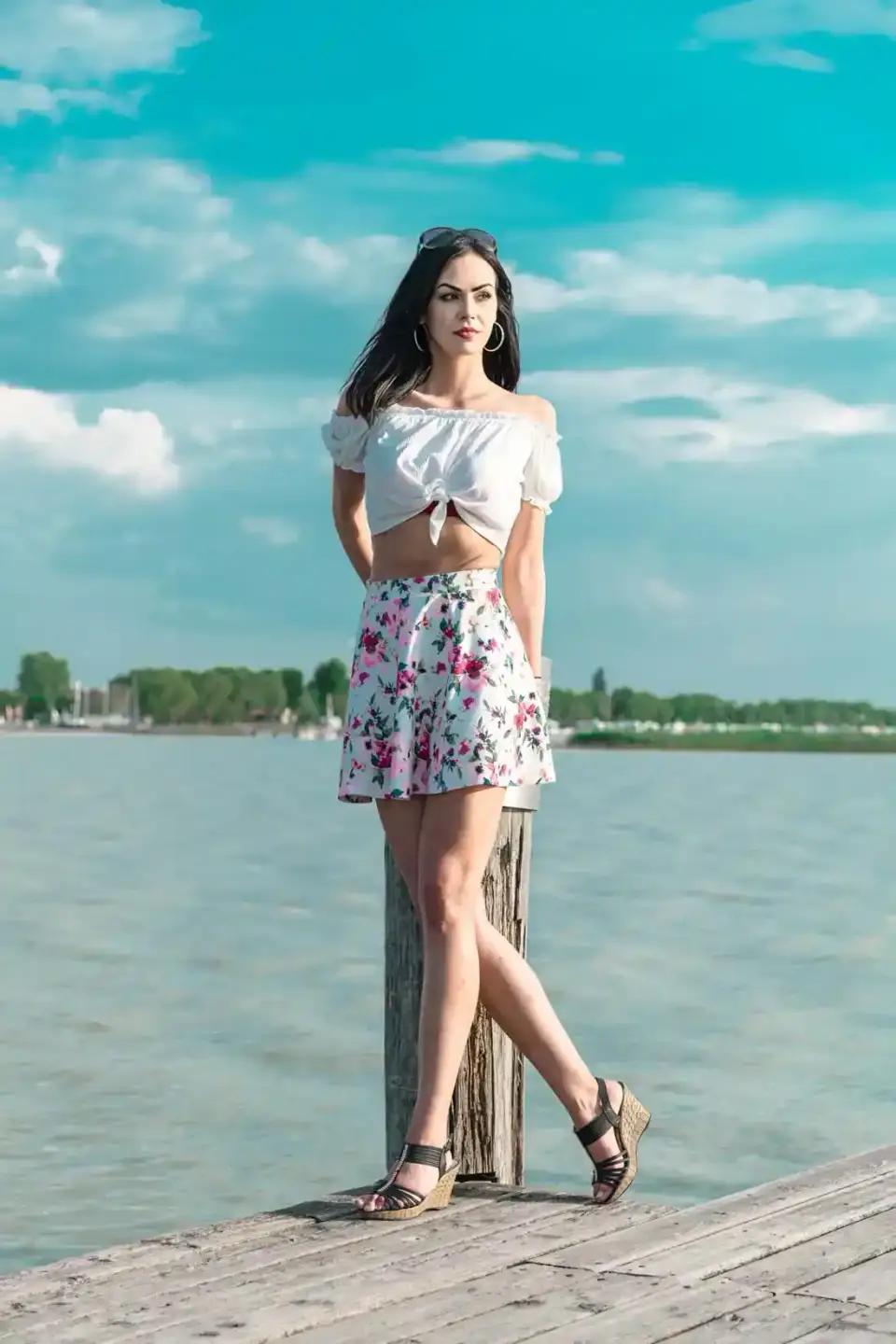 Junge Frau in weißem Crop-Top und floralem Rock steht auf einem Holzsteg am Wasser, Portrait Wien vor malerischer Seenlandsch