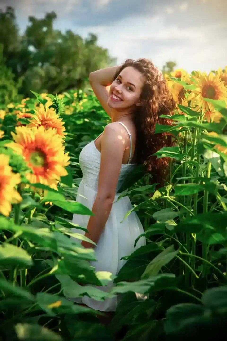Junge Frau in weißem Kleid posiert lächelnd in einem Sonnenblumenfeld – professionelles Portrait von Portraitfotograf Wien