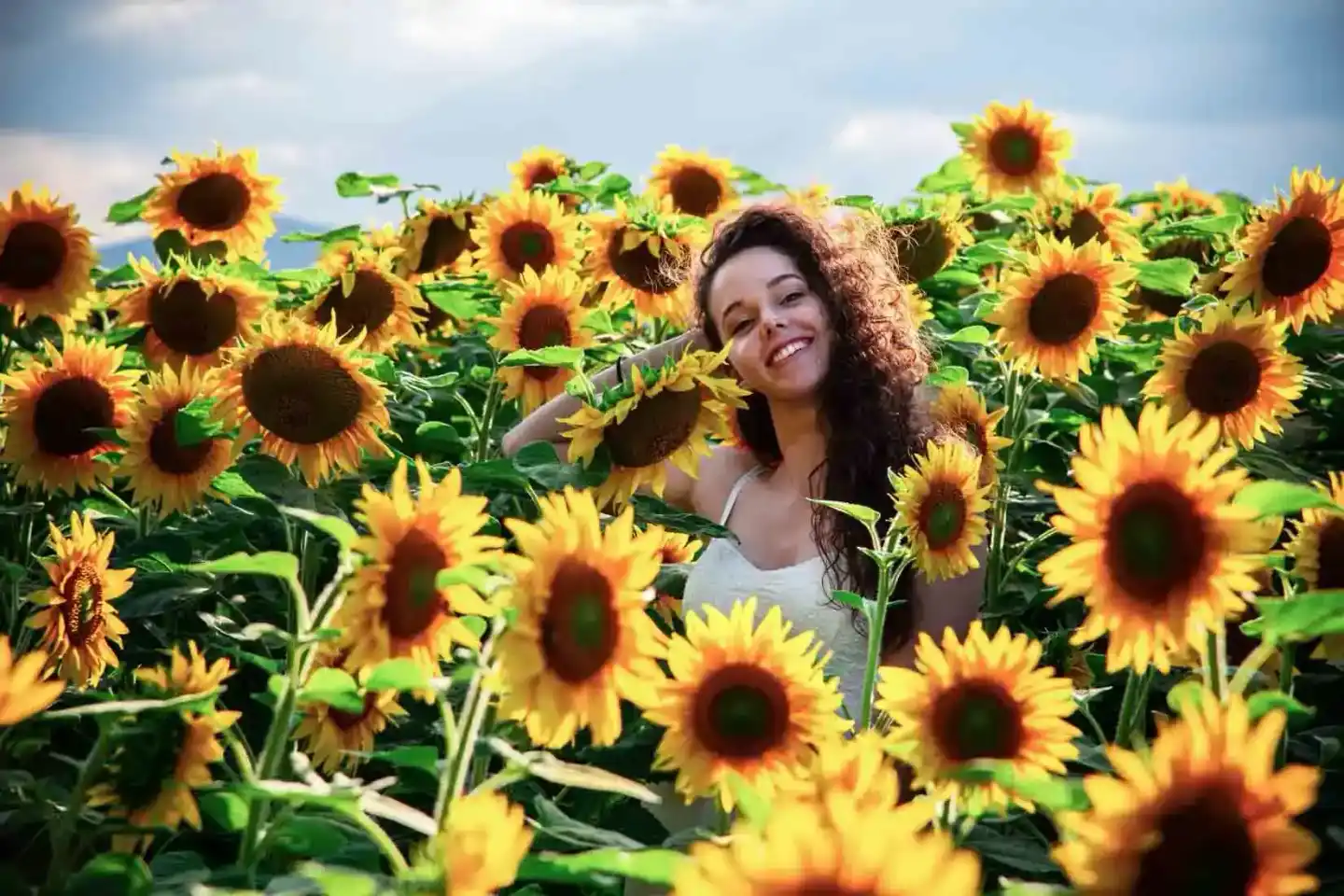 Junge Frau mit langen dunklen Haaren lächelt inmitten eines Sonnenblumenfeldes während eines professionellen Fotoshootings in