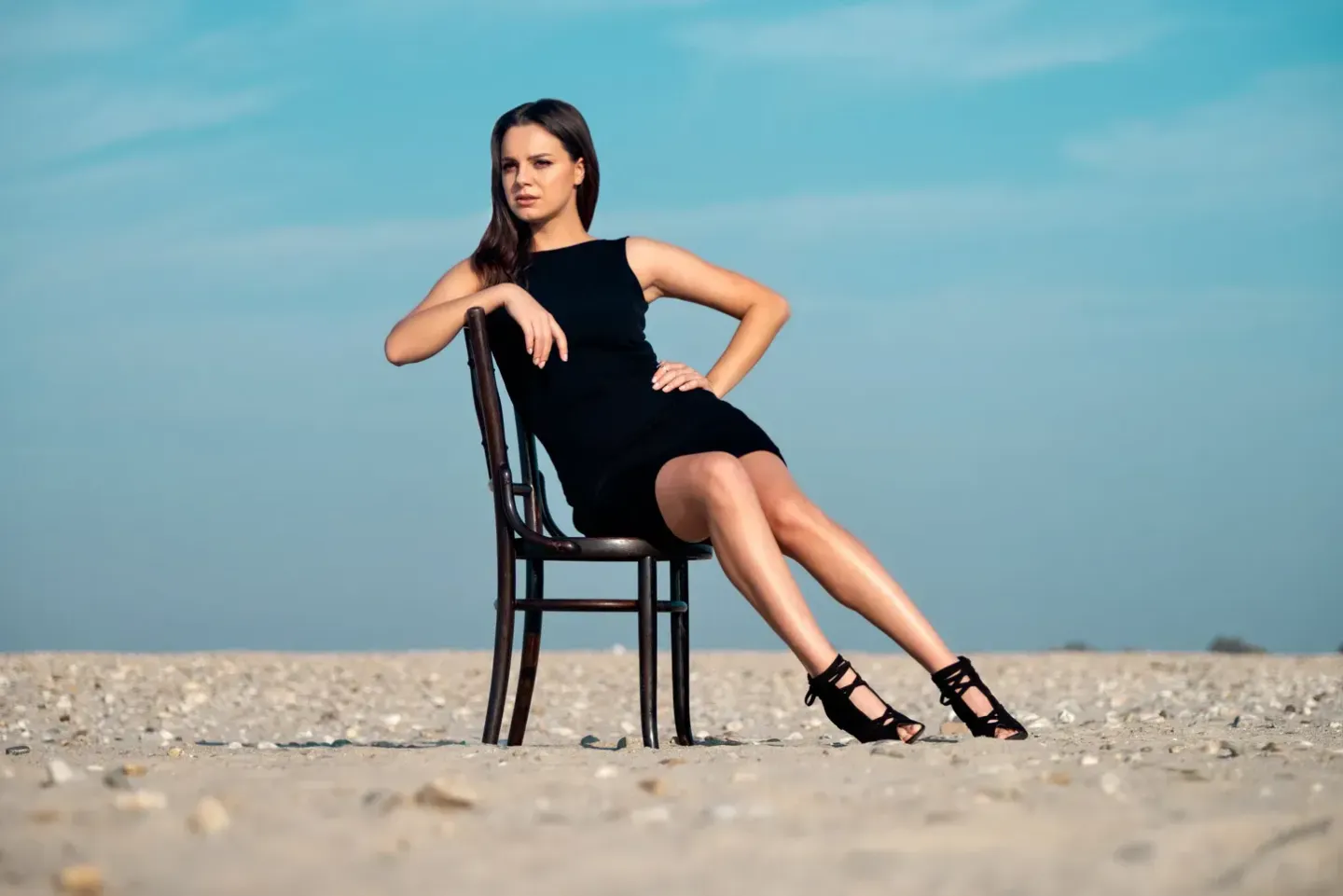 Frau sitzt auf Stuhl am Strand im schwarzen Kleid, professionelles Portrait Wien mit Meereshintergrund
