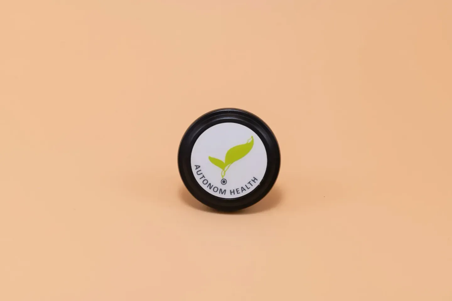 Schwarze runde Dose mit grünem Blatt-Logo und Aufschrift AUTONOM HEALTH auf beigem Hintergrund – Produktfotografie Wien