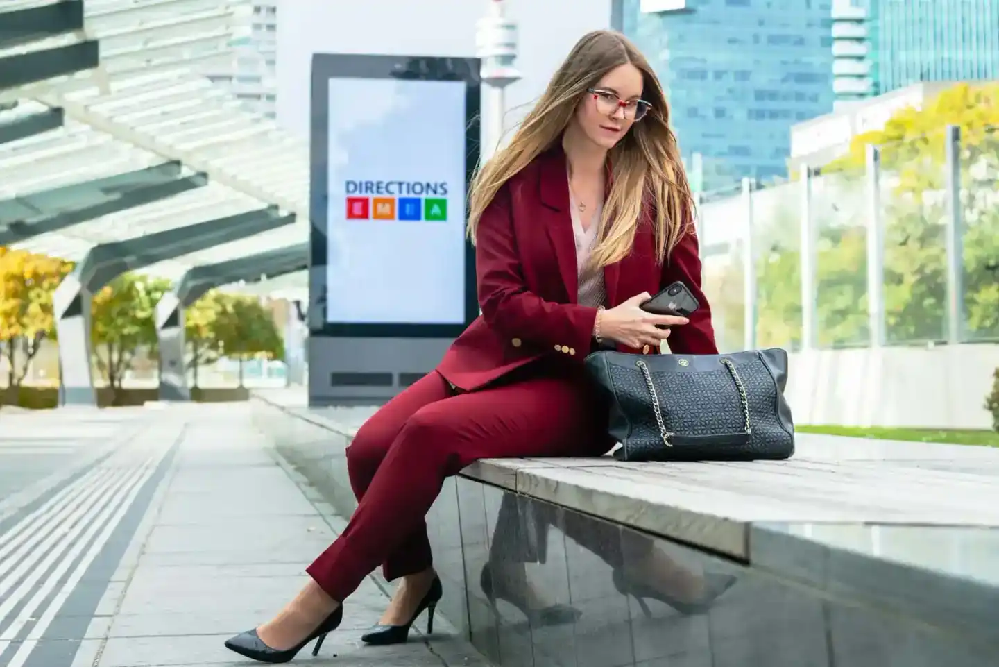 Geschäftsfrau in burgunderrotem Anzug sitzt auf einer Bank mit schwarzer Designer-Tasche, im Hintergrund moderne Architektur 