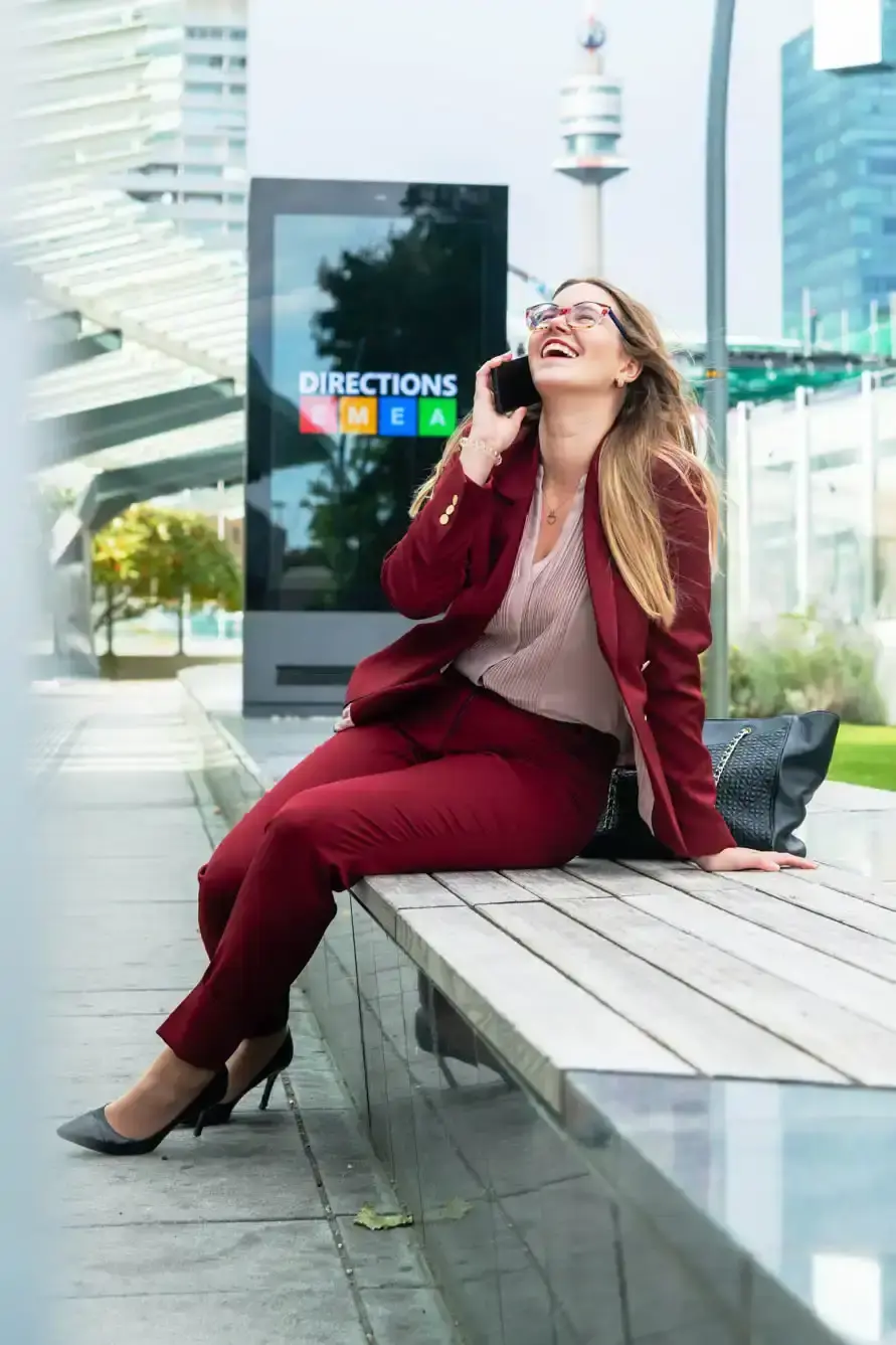 Frau in eleganter Burgunderanzug sitzt auf einer Bank vor einem Directions-Display in Wien, lacht beim Telefonieren