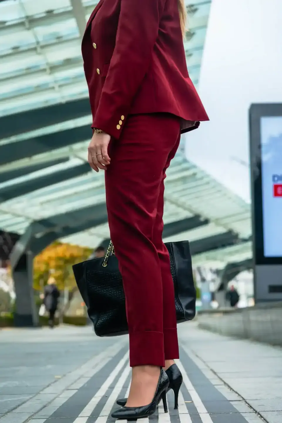 Frau in burgunderrotem Blazer und passender Hose mit schwarzer Handtasche und High Heels vor modernem Gebäude – Produktfotogr