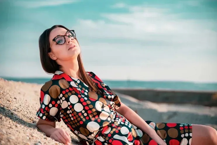 Frau in farbenfrohem Kleid mit Muster sitzt am Strand, trägt Sonnenbrillen – Beispiel für professionelle Produktfotografie Wi