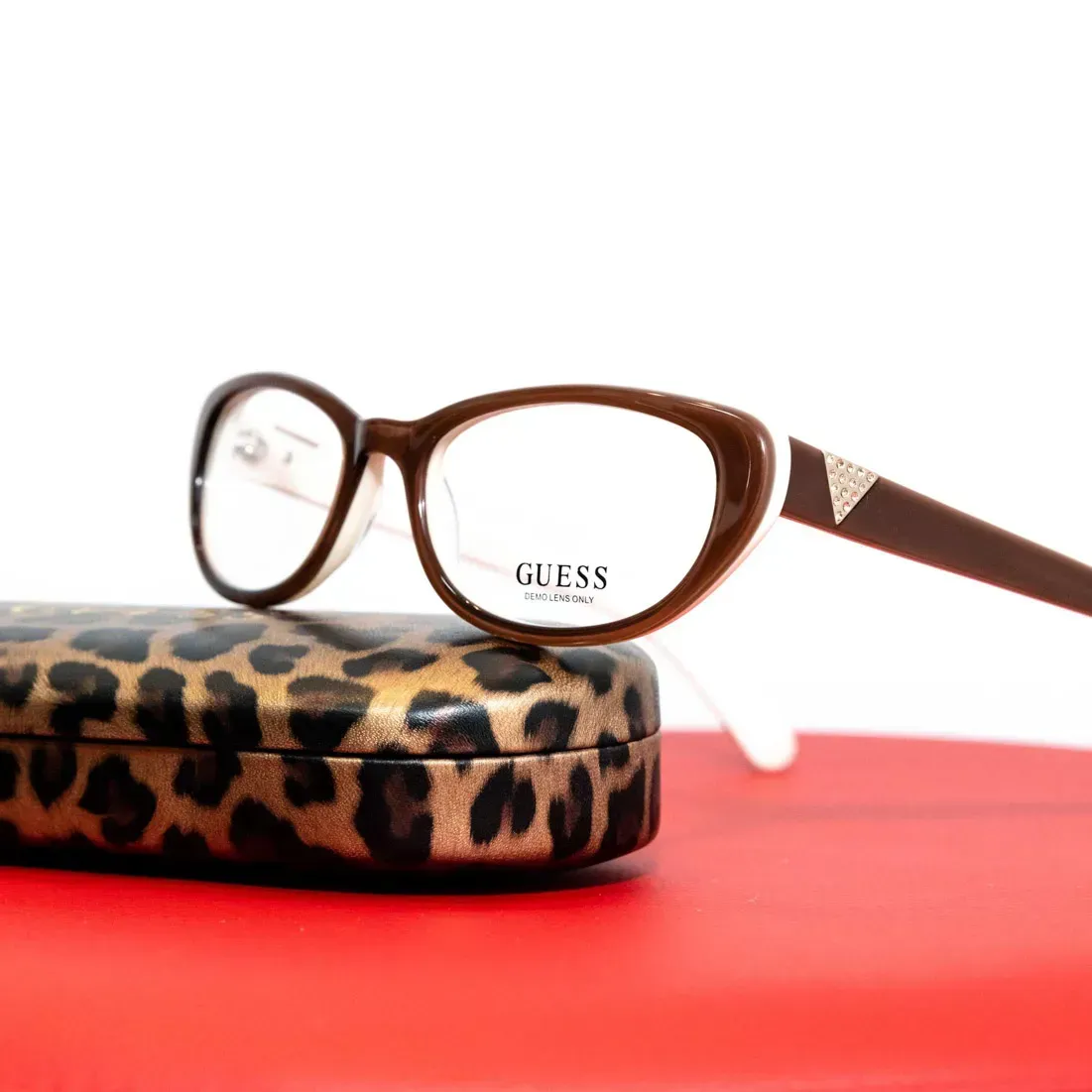 Braune GUESS Brille mit Leoparden-Muster Etui auf rotem Hintergrund – Produktfotografie Wien
