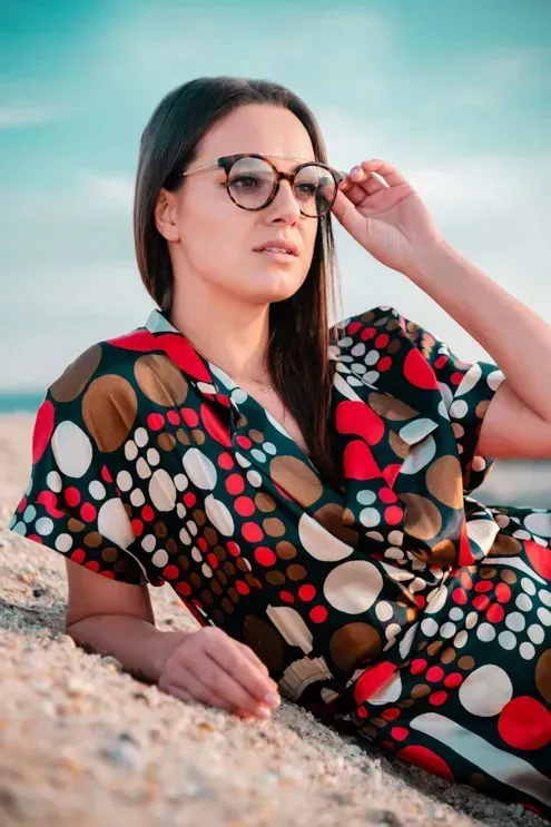 Junge Frau mit Brille in gemustertem Kleid sitzt am Strand – Produktfotografie Wien