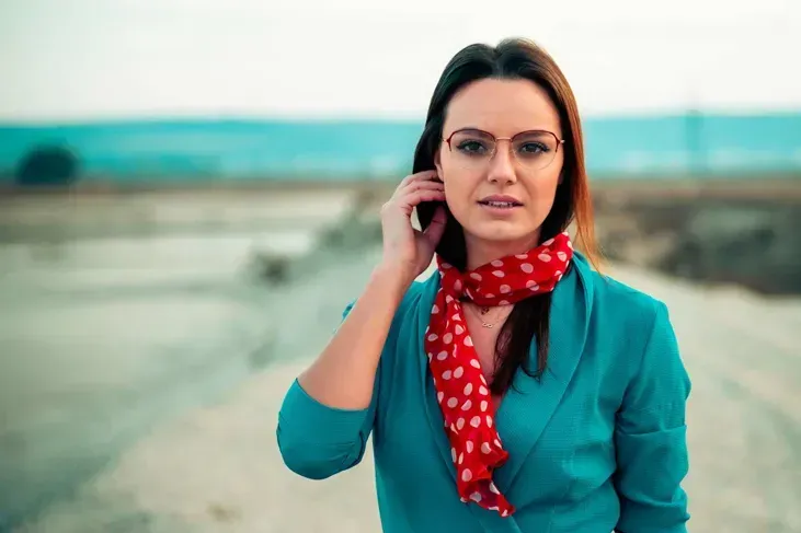 Frau mit Brille in türkisblauem Blazer und rotem Schal telefoniert in ländlicher Landschaft – Werbefotografie Wien