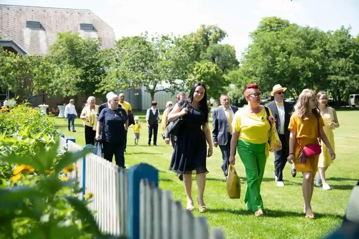 Hochzeitsgäste spazieren über eine grüne Wiese mit Blumendekorationen und historischem Gebäude im Hintergrund