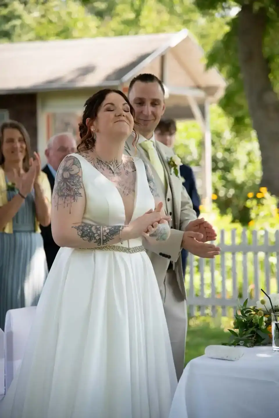 Brautpaar bei der Trauung im Garten: Braut im weißen Kleid mit Tattoos und Bräutigam im beigen Anzug, Hochzeitsfotograf Wien