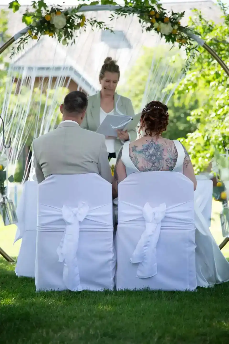 Brautpaar sitzt unter einer grünen Trauungszeremonie-Arch, Hochzeitsfotograf dokumentiert den emotionalen Moment im Freien