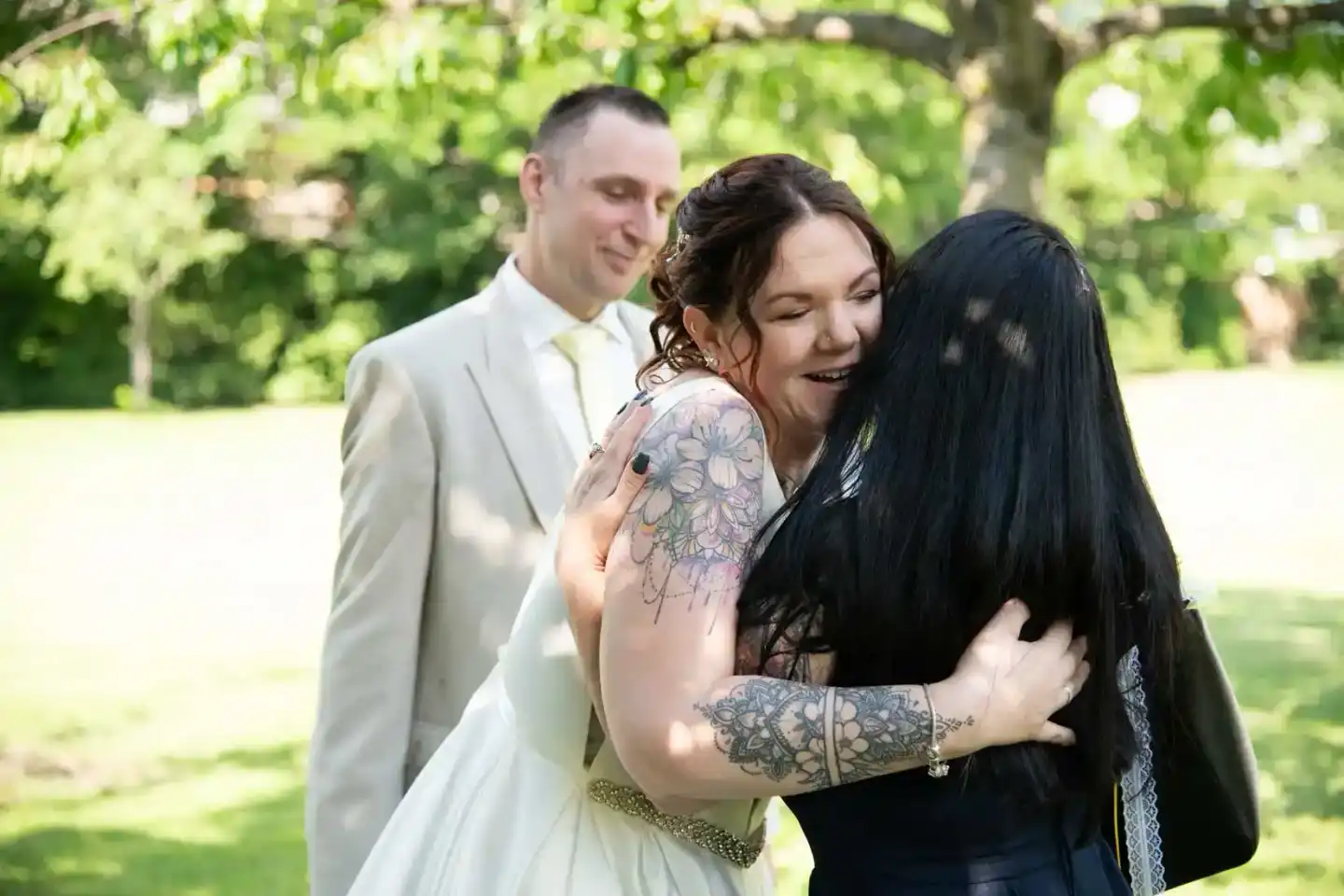 Braut in weißem Kleid umarmt Hochzeitsgast, Bräutigam im Hintergrund, grüner Garten – Hochzeitsfotografie Wien