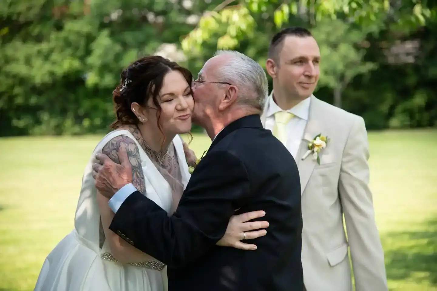 Braut mit Tattoo umarmt ihren Vater, während der Bräutigam zusieht – emotionaler Moment bei einer Hochzeit in Wien