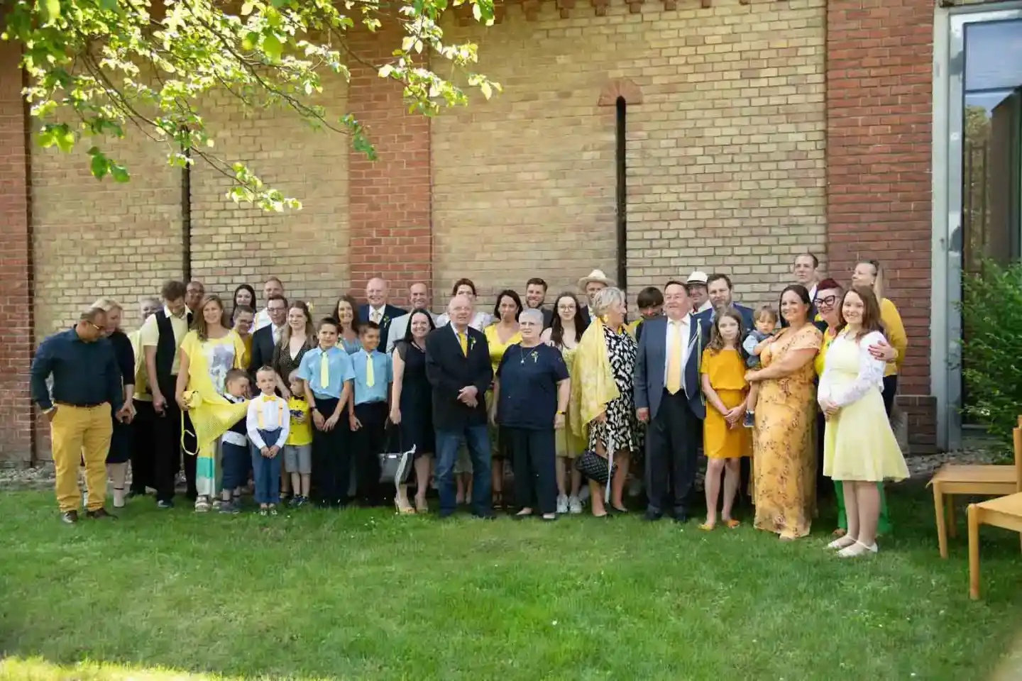 Große Hochzeitsgesellschaft mit Familie und Gästen in gelben und schwarzen Outfits vor einer Backsteinmauer, fotografiert von