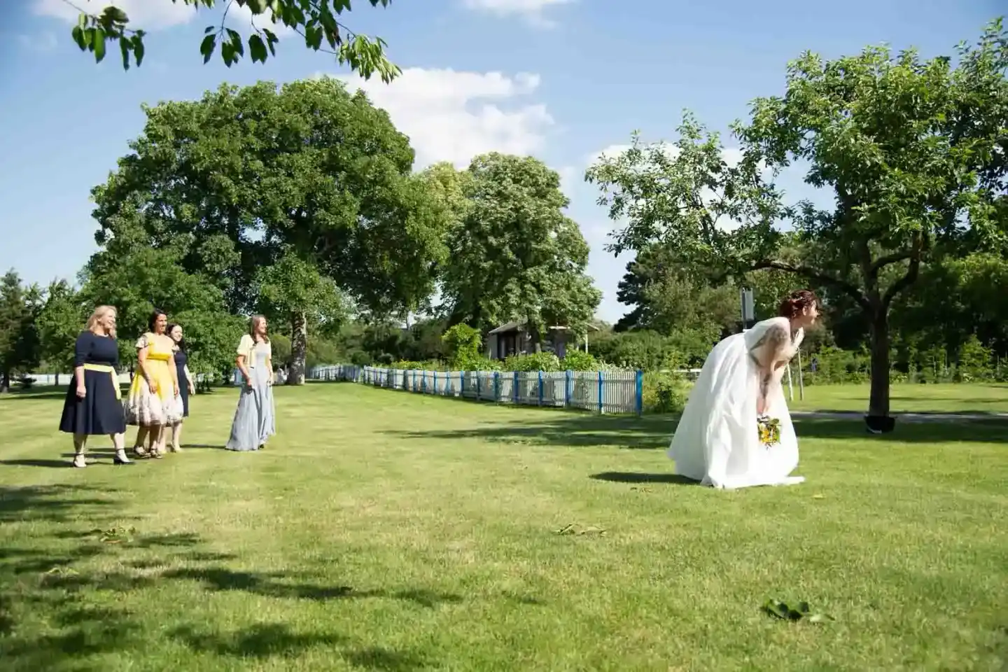 Braut in weißem Kleid beugt sich auf grüner Wiese, während Hochzeitsgäste beobachten – Hochzeitsfotografie Wien