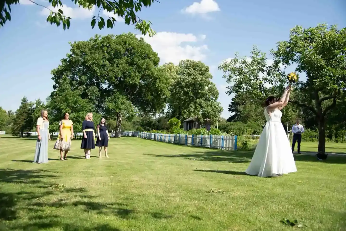 Braut in weißem Hochzeitskleid wirft Brautstrauß auf einer grünen Wiese, Hochzeitsgäste beobachten das Bouquet-Toss