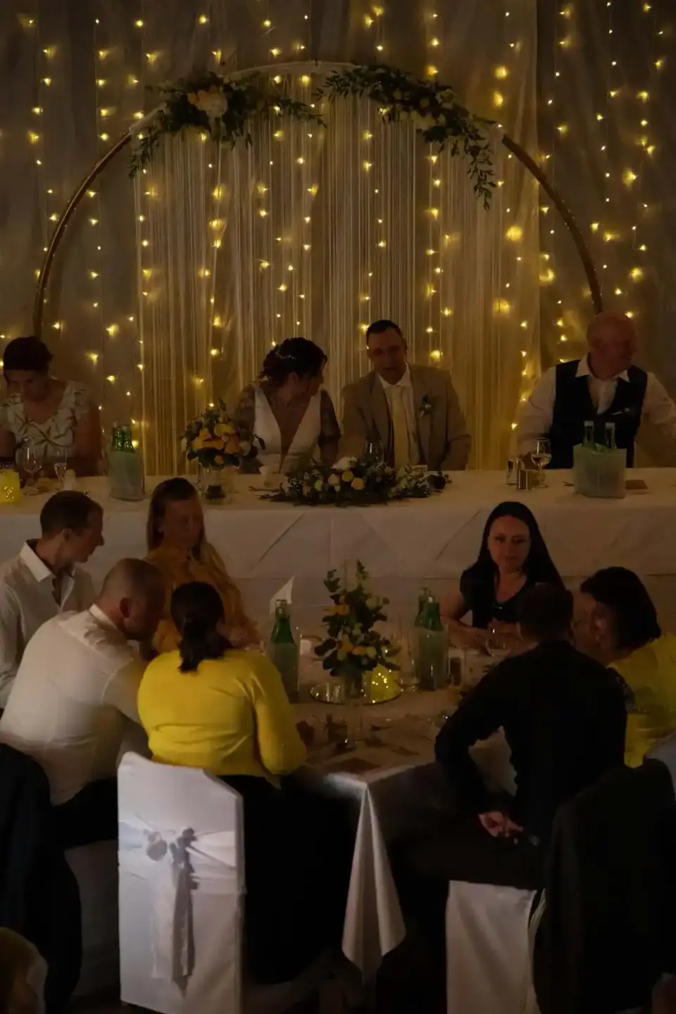 Hochzeitsfotografie Wien: Brautpaar und Gäste beim Hochzeitsessen unter leuchtender Lichterkette und Blumenbogen