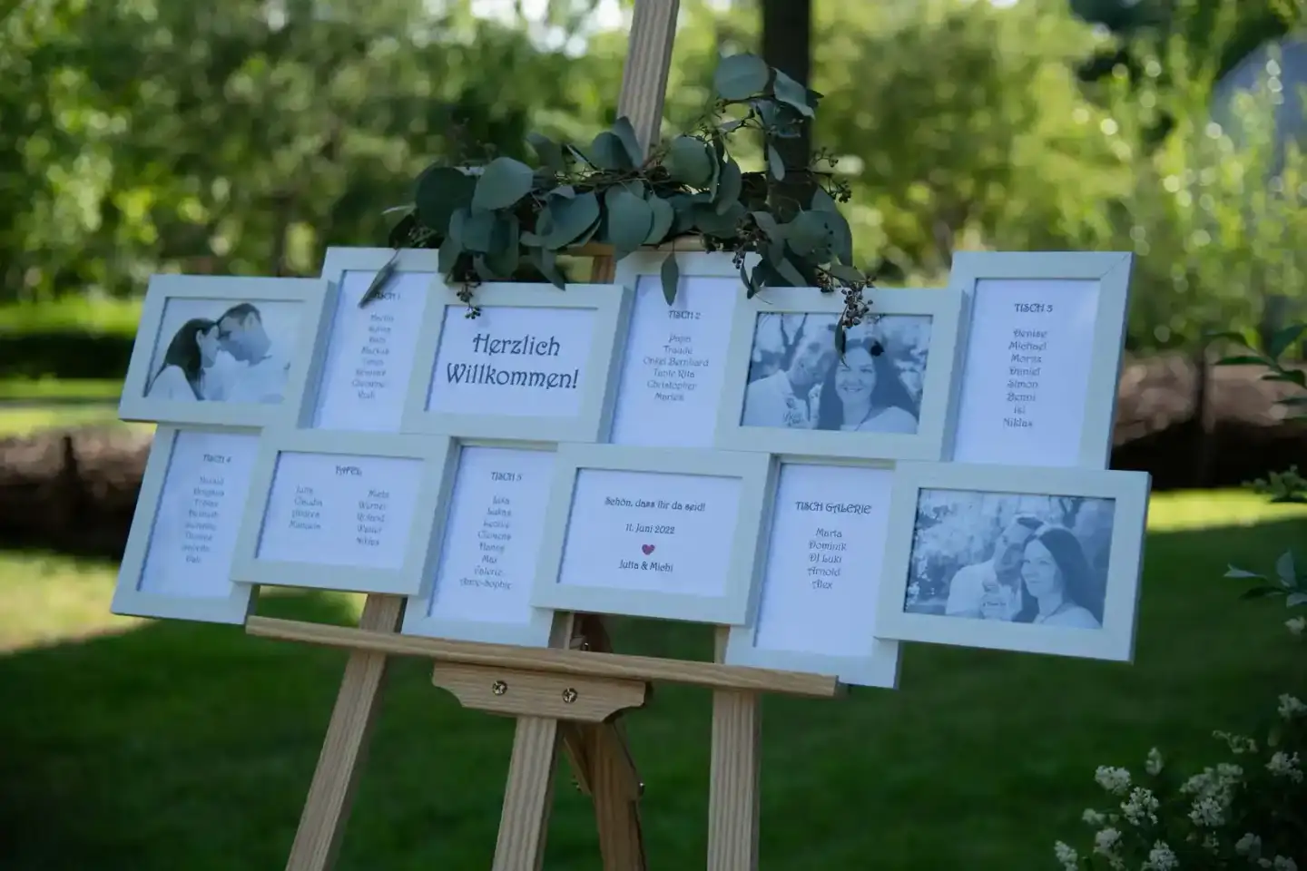 Willkommenstafel zur Hochzeit mit Fotos und Gästelisten, dekoriert mit Eukalyptus auf grünem Rasen