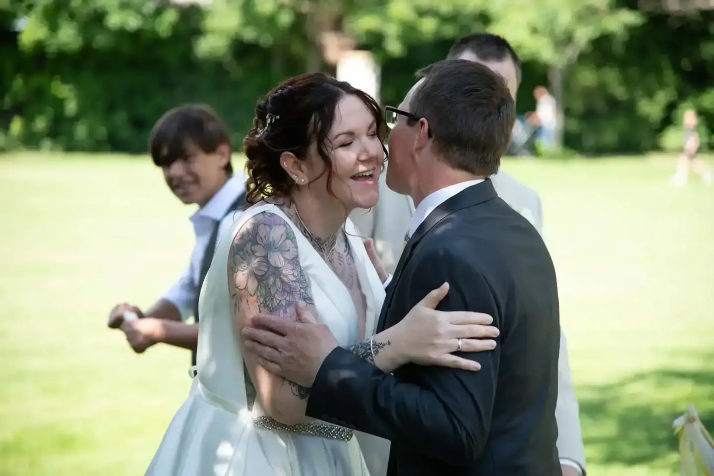 Brautpaar bei der Hochzeitszeremonie im grünen Park, Bräutigam umarmt lachende Braut mit Tattoo